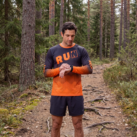 Male runner wearing a black and orange RUN REST LIFT REPEAT long sleeve shirt on a wooded trail looking at his watch