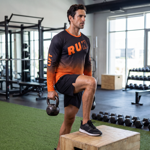 Male athlete wearing a black and orange RUN REST LIFT REPEAT long sleeve shirt while doing weighted step-ups in a gym