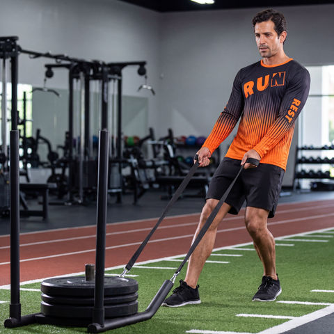 Male athlete pulling a weighted sled while wearing a black and orange RUN REST LIFT REPEAT long sleeve performance shirt in a gym