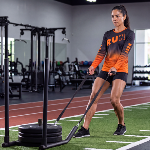 Female athlete pulling a weighted sled while wearing a black and orange RUN REST LIFT REPEAT long sleeve performance shirt