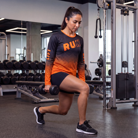 Female athlete doing weighted lunges in a gym while wearing a black and orange RUN REST LIFT REPEAT long sleeve performance shirt